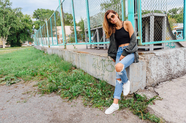 Fashion portrait of trendy young woman wearing sunglasses, jeans with halls and bomber jacket in the city. Copy space. - Fotografie, Obrázek