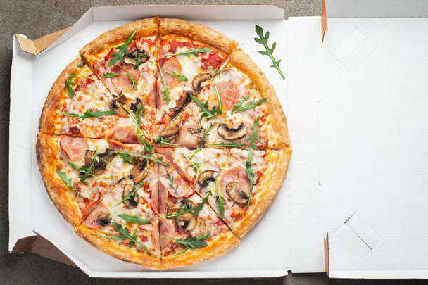Italiaanse fastfood. Heerlijke hete pizza in een doos met ham, champignons gesneden en geserveerd op witte tafel, close-up. Concept van snelle en ongezonde voeding. - Foto, afbeelding