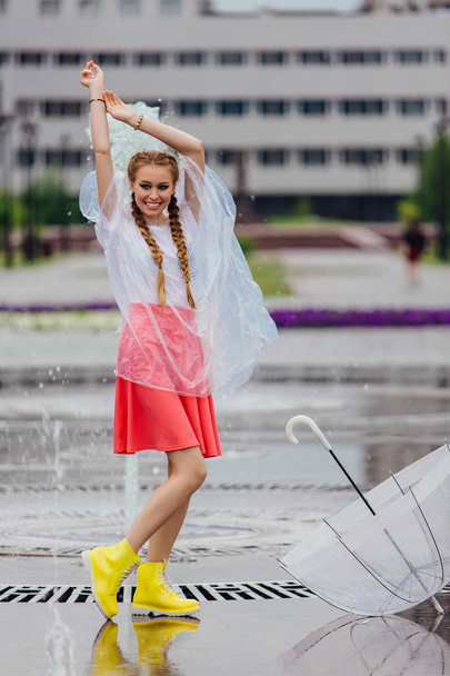 Young pretty girl with two braids in yellow boots and with transparent umbrella stands near fountain. Rainy day in city. - Фото, зображення