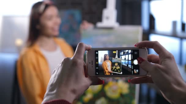 Close-up vrienden handen nemen foto's van Aziatische vrouw schilder lachend met haar bloemen schilderij in huis van kunststudio op mobiele telefoon scherm. Wazig gelukkig kunstenaar poseren met haar schilderij. - Video