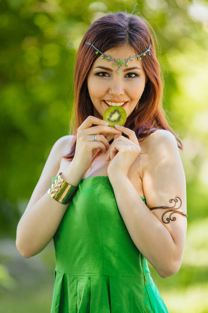 Porträt einer schönen jungen Asiatin in grünem Kleid und Diadema auf dem Kopf, die in Kiwi beißt - Foto, Bild