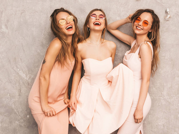Τρία μικρά όμορφα χαμογελώντας hipster κορίτσια σε μοντέρνο καλοκαιρινό φως ροζ φορέματα. Σέξι ανέμελη γυναίκες ποζάρουν κοντά γκρίζο τοίχο. Θετικά πρότυπα σε στρογγυλά γυαλιά ηλίου διασκεδάζοντας - Φωτογραφία, εικόνα