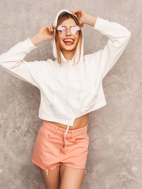 Retrato de jovem bela menina sorridente na moda verão hipster roupas esportivas. Mulher despreocupada Sexy posando perto da parede cinza. Modelo positivo se divertindo em óculos de sol
  - Foto, Imagem