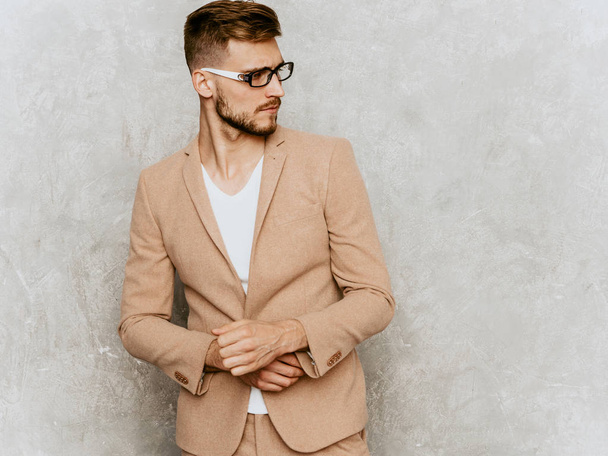 Portrait of handsome smiling hipster lumbersexual businessman model wearing casual beige suit. Fashion stylish man posing against gray wall in spectacles - 写真・画像