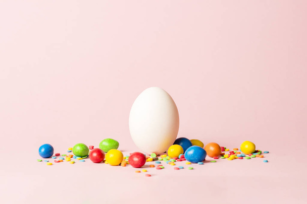 Huevo de Pascua blanco y dulces multicolores sobre un fondo rosa. Concepto de celebración Pascua
 - Foto, Imagen