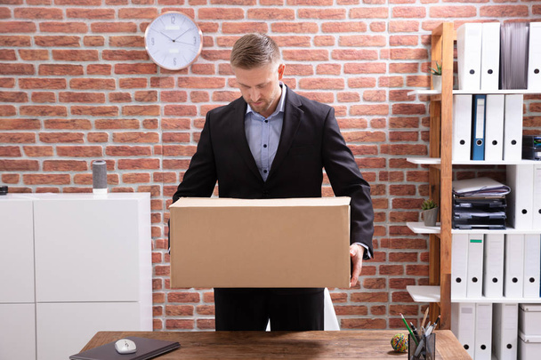 Young Businessman Packing His Belongings In Cardboard Box At Workplace - Фото, изображение