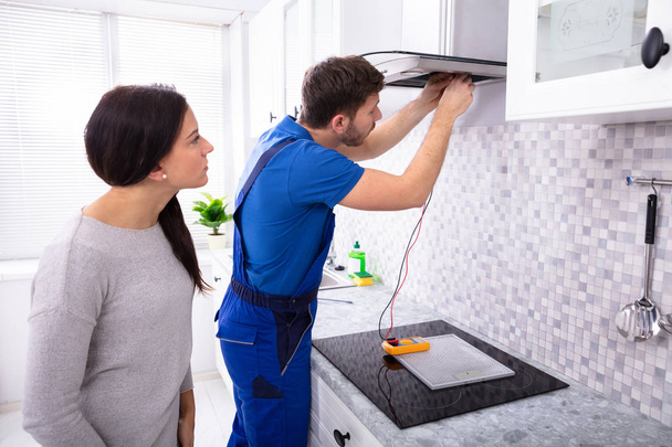 Male Repairman Checking Extractor Filter With Digital Multimeter While Woman Standing In Kitchen - Photo, Image