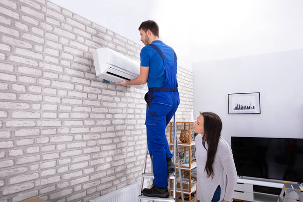 Young Woman Looking At Male Technician Fixing Air Conditioner On Brick Wall - Foto, imagen
