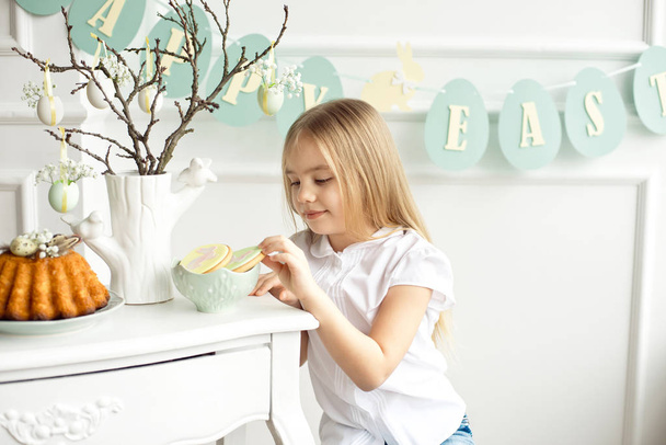 A cute toddler girl in a white shirt is eating with Easter eggs on Easter day. - Foto, Imagen