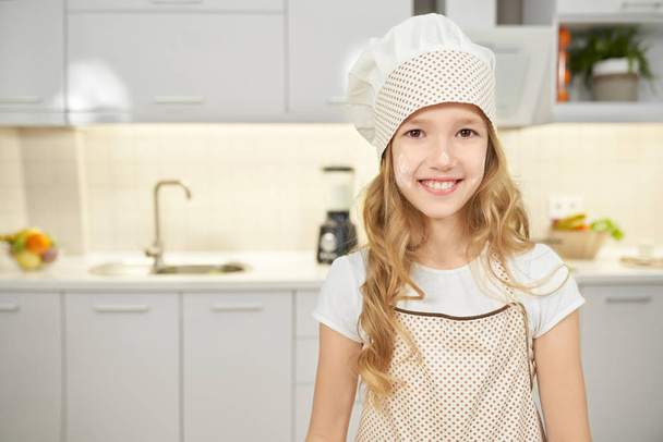 Happy girl in apron and chef hat posing in kitchen. - Zdjęcie, obraz