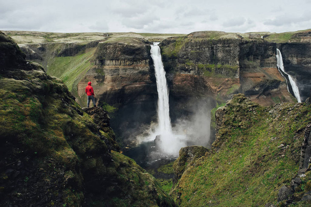 Man in rode jas op zoek op adembenemende IJslandse waterval  - Foto, afbeelding