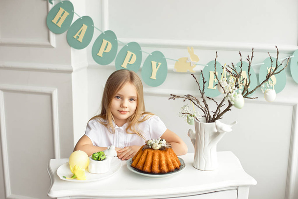 Cute little blonde girl sits behind a white vintage table enjoys a cooked easter cupcake - Fotoğraf, Görsel