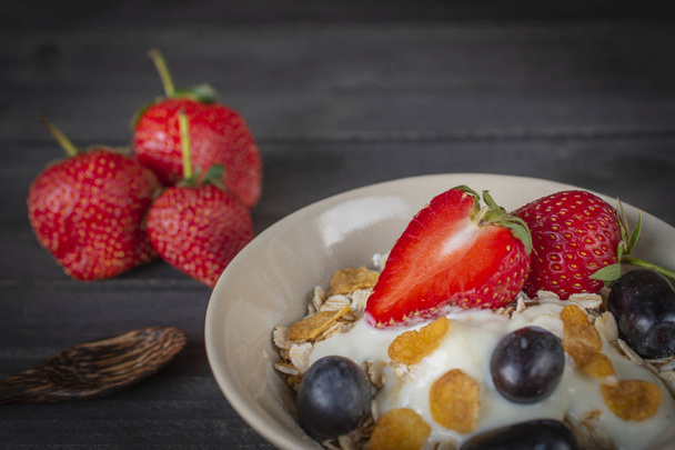Yoghurt mix oatmeal, strawberry and grape topping in white bowl on the wood table with spoon and strawberries placed beside. - Photo, Image