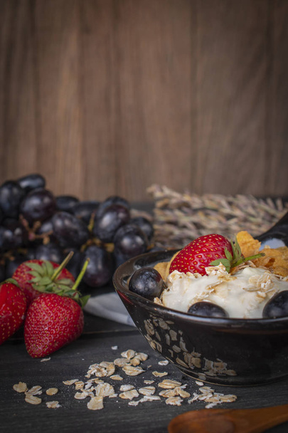 Yoghurt mix havermout, aardbeien en druiven topping in zwarte kom op houten tafel met blauw en wit gestreepte stof, lepel, aardbei, druif en havermout geplaatst rond. - Foto, afbeelding