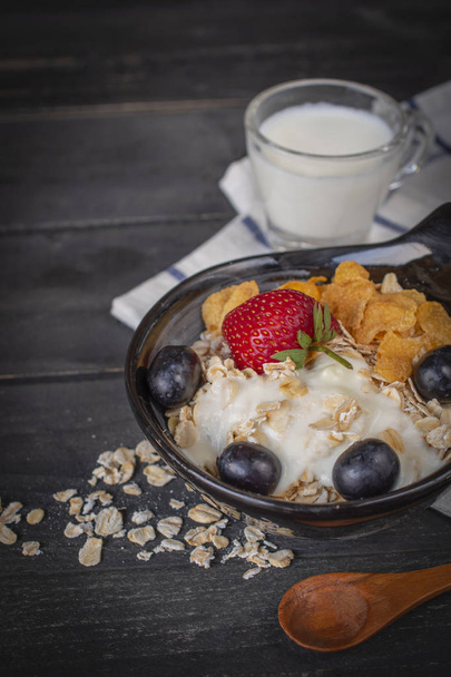 Yoghurt mix havermout, aardbeien en druiven topping in zwarte kom op houten tafel met blauw en wit gestreepte stof, lepel, havermout en melk in glas geplaatst rond. - Foto, afbeelding