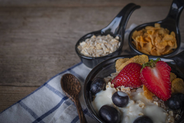 Joghurt-Mix Haferflocken, Erdbeeren und Trauben in schwarzer Schüssel auf blau-weiß gestreiftem Stoff und Holztisch mit Löffel, Cornflakes und Haferflocken. - Foto, Bild