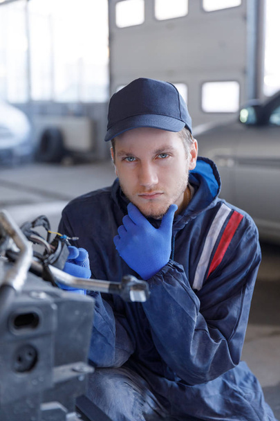 Young mechanic in the auto repair shop. Thumbs up.A young mechanic in an auto repair shop, folded his arms in front of him. The car in the background is covered with a cover. - Photo, image