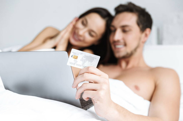 Beautiful young couple laying in bed, shopping with laptop and credit card - Foto, Imagem