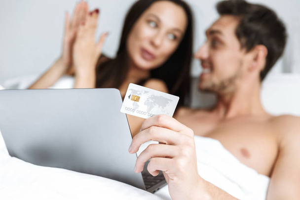 Beautiful young couple laying in bed, shopping with laptop and credit card - Foto, afbeelding