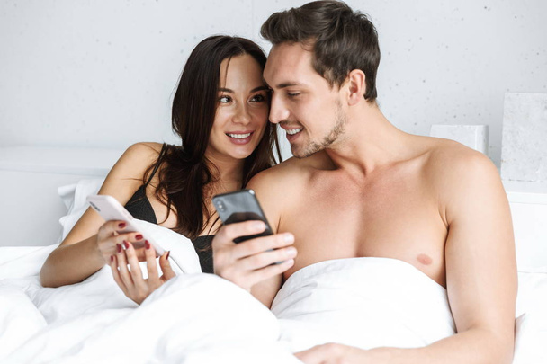 Beautiful young couple laying in bed, holding mobile phones - 写真・画像