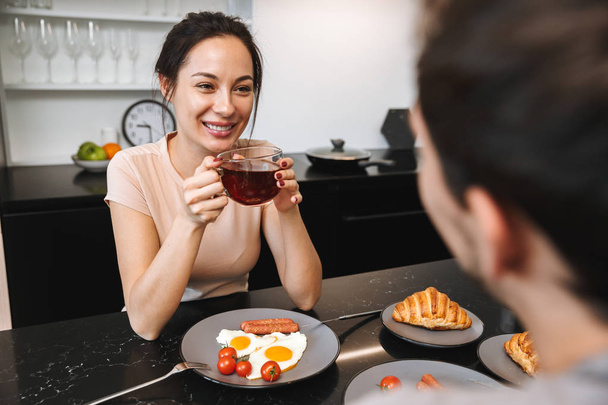 Image of a young happy loving couple indoors in home at kitchen have a breakfast drinking tea. - Foto, Imagen