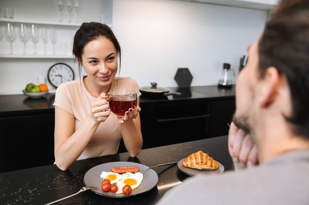Image of a young happy loving couple indoors in home at kitchen have a breakfast drinking tea. - Photo, image