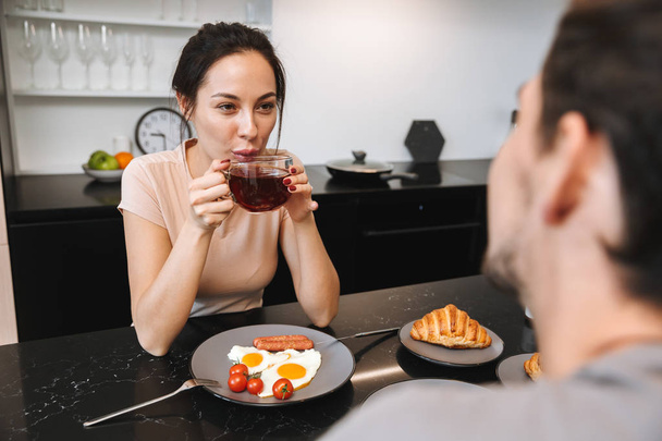 Image of a young happy loving couple indoors in home at kitchen have a breakfast drinking tea. - 写真・画像