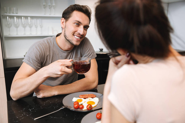 Image of a young happy loving couple indoors in home at kitchen have a breakfast drinking tea. - Photo, Image