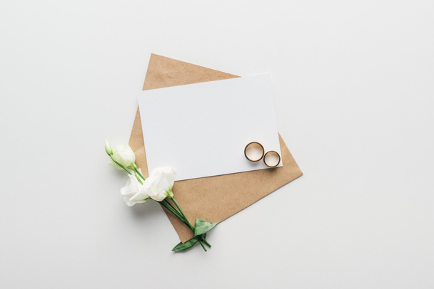 top view of envelope with empty card, flowers and golden wedding rings on grey background - Фото, изображение