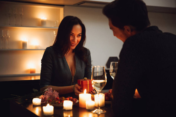 Beautiful passionate couple having a romantic candlelight dinner at home, drinking wine, woman getting a gift box - Фото, изображение