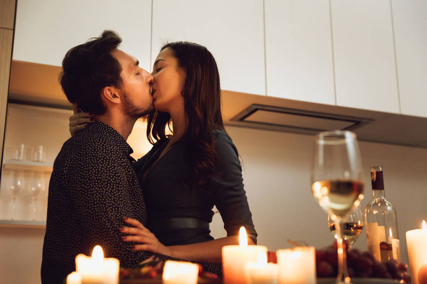 Beautiful passionate couple having a romantic candlelight dinner at home, kissing - Фото, изображение
