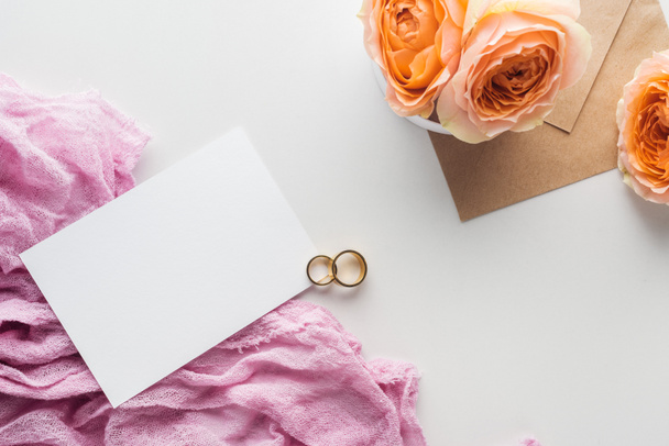 top view of empty card, envelope, flowers, cloth and golden wedding rings on grey background - 写真・画像
