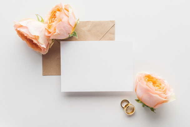 top view of empty card over brown craft paper envelope, roses and golden wedding rings on grey background - Fotografie, Obrázek