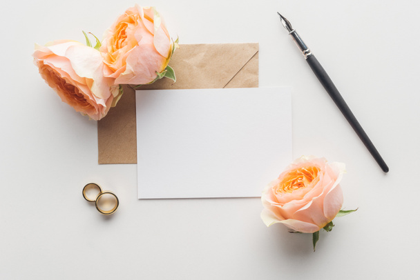 Draufsicht auf leere Karte mit braunem Bastelpapier-Umschlag, Tuschstift, Blumen und Trauringen auf grauem Hintergrund - Foto, Bild
