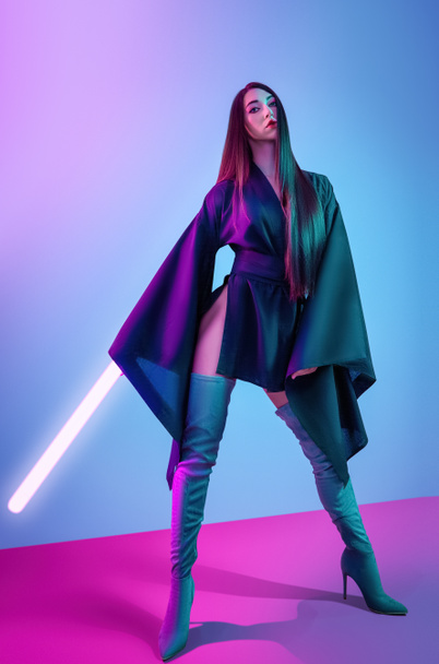 Concept on cosmic cosplay. Contemporary portrait a young athletic woman in traditional Japanese black kimono, an Asian hat and highboots is holding a lightsaber and posing on neon blue-pink background - Фото, изображение