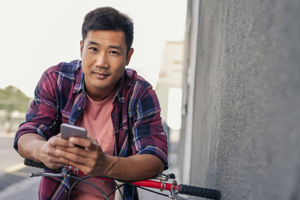 Portrait of a smiling young Asian man in a plaid shirt leaning on his bike on a city sidewalk sending a text on his cellphone - 写真・画像