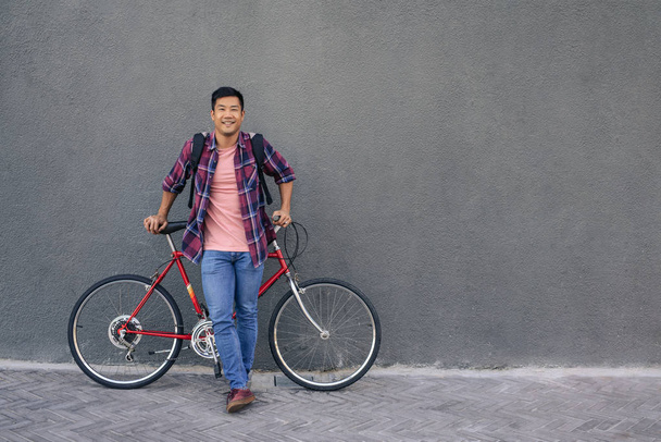Portrait of a smiling young Asian man in a plaid shirt standing with his bike in front of a gray wall in the city - Φωτογραφία, εικόνα