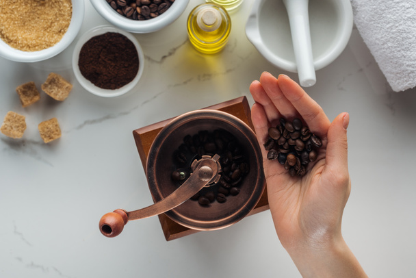 vista parcial de la mano femenina con granos de café, molino de mano, agarradera y diferentes ingredientes para la fabricación de cosméticos en la superficie blanca
 - Foto, imagen