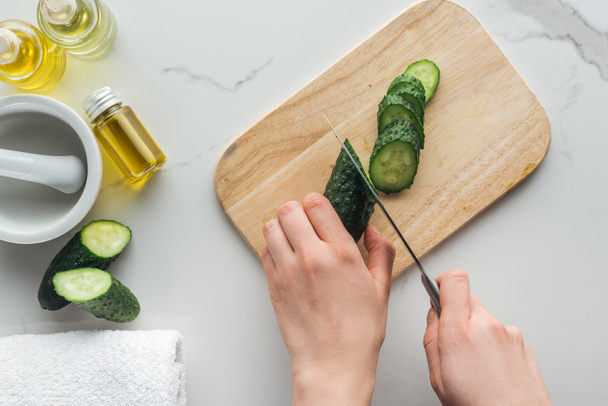 cropped view of woman cutting cucumber on wooden cutting desk, pounder and various cosmetic ingredients on white surface - Photo, Image