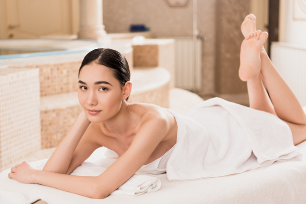 привлекательная азиатка в полотенце лежит на массажном столе и смотрит в камеру
  - Фото, изображение