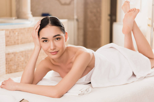 beautiful asian woman in towel lying on massage table and looking at camera  - Zdjęcie, obraz