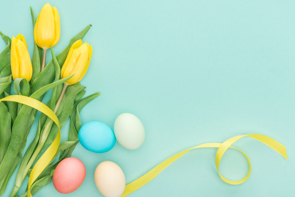 top view of yellow tulip flowers with ribbon and easter eggs isolated on blue with copy space - Foto, afbeelding