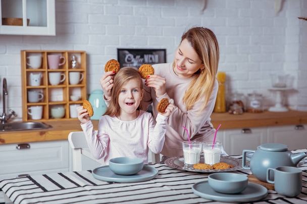 fröhliches kleines Mädchen und ihre schöne junge Mutter frühstücken zusammen in einer weißen Küche. Sie haben Spaß und spielen mit Keksen. Mütterfürsorge und Liebe. - Foto, Bild