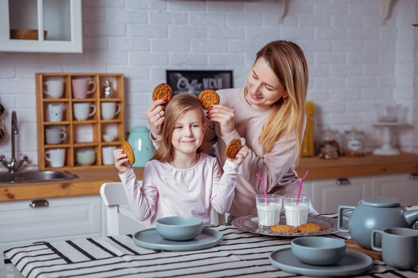 Happy little girl and her beautiful young mother have breakfast together in a white kitchen. They are having fun and playing with cookies. Maternal care and love. - Photo, Image