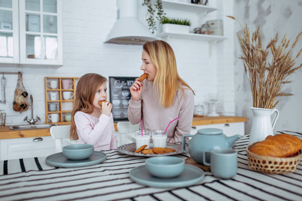 Happy little girl and her beautiful young mother have breakfast together in a white kitchen. They drink milk and eat cookies. Maternal care and love. Horizontal photo. - Foto, Imagem