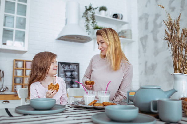 Happy little girl and her beautiful young mother have breakfast together in a white kitchen. They drink milk and eat cookies. Maternal care and love. Horizontal photo. - Photo, Image