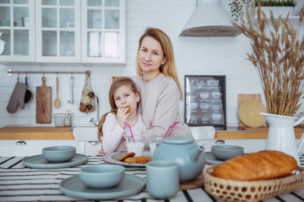 Happy little girl and her beautiful young mother have breakfast together in a white kitchen. Mom hugs daughter. Maternal care and love. Horizontal photo. - 写真・画像