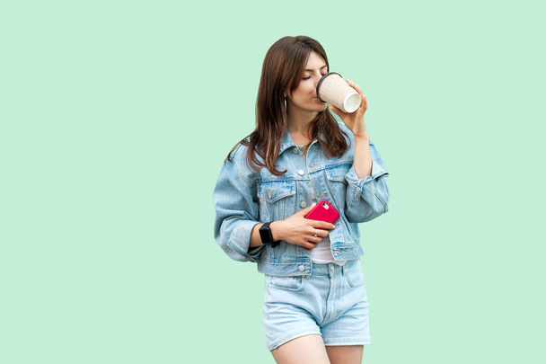 beautiful young brunette woman in casual denim style holding smartphone and drinking from disposable cup on green background. - Foto, Imagen