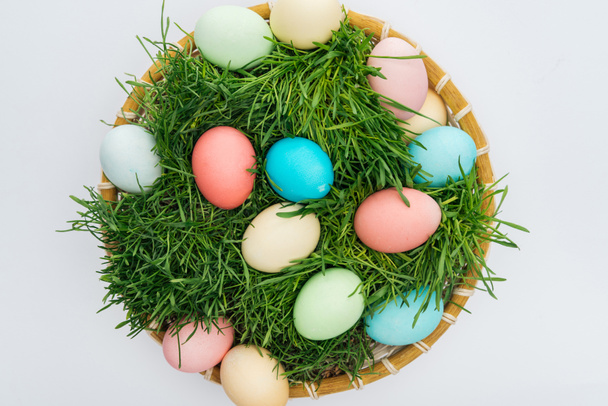 top view of colorful easter eggs in wicker plate with grass isolated on white - Foto, imagen