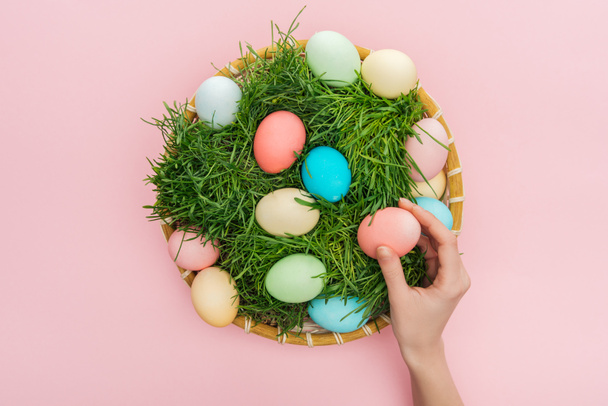 cropped view of female hand with pastel easter eggs in wicker plate with grass isolated on pink  - Valokuva, kuva